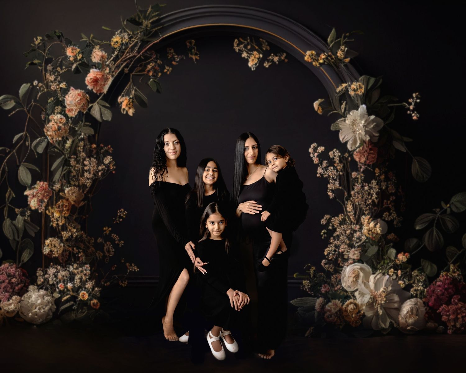 Family portrait of five women in black dresses posed before a floral arch with dark moody backdrop.