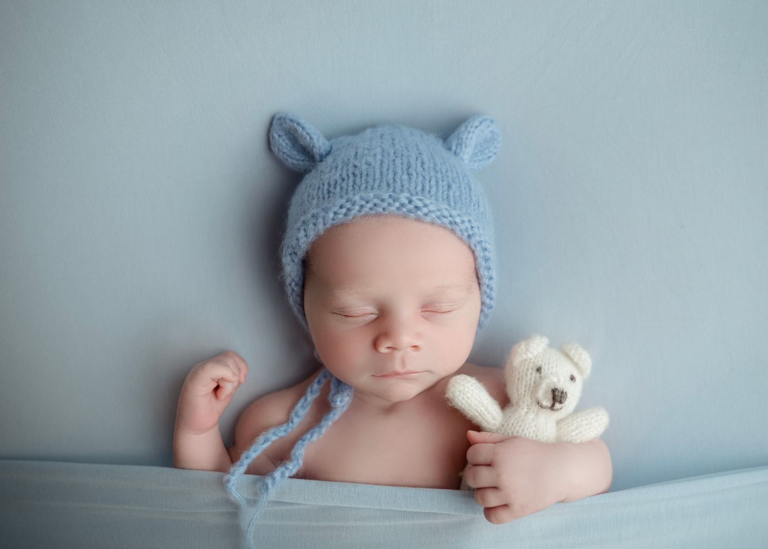 Sleeping newborn baby wearing a blue knit bear bonnet hat, holding a small white teddy bear toy.