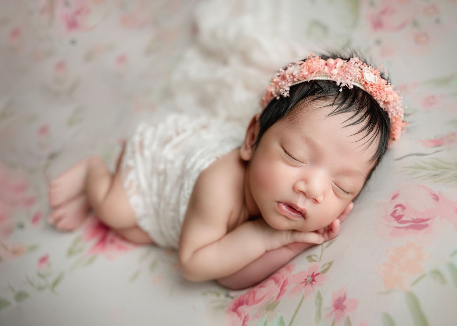 Sleeping newborn baby girl wearing a pink floral headband on a soft floral backdrop.
