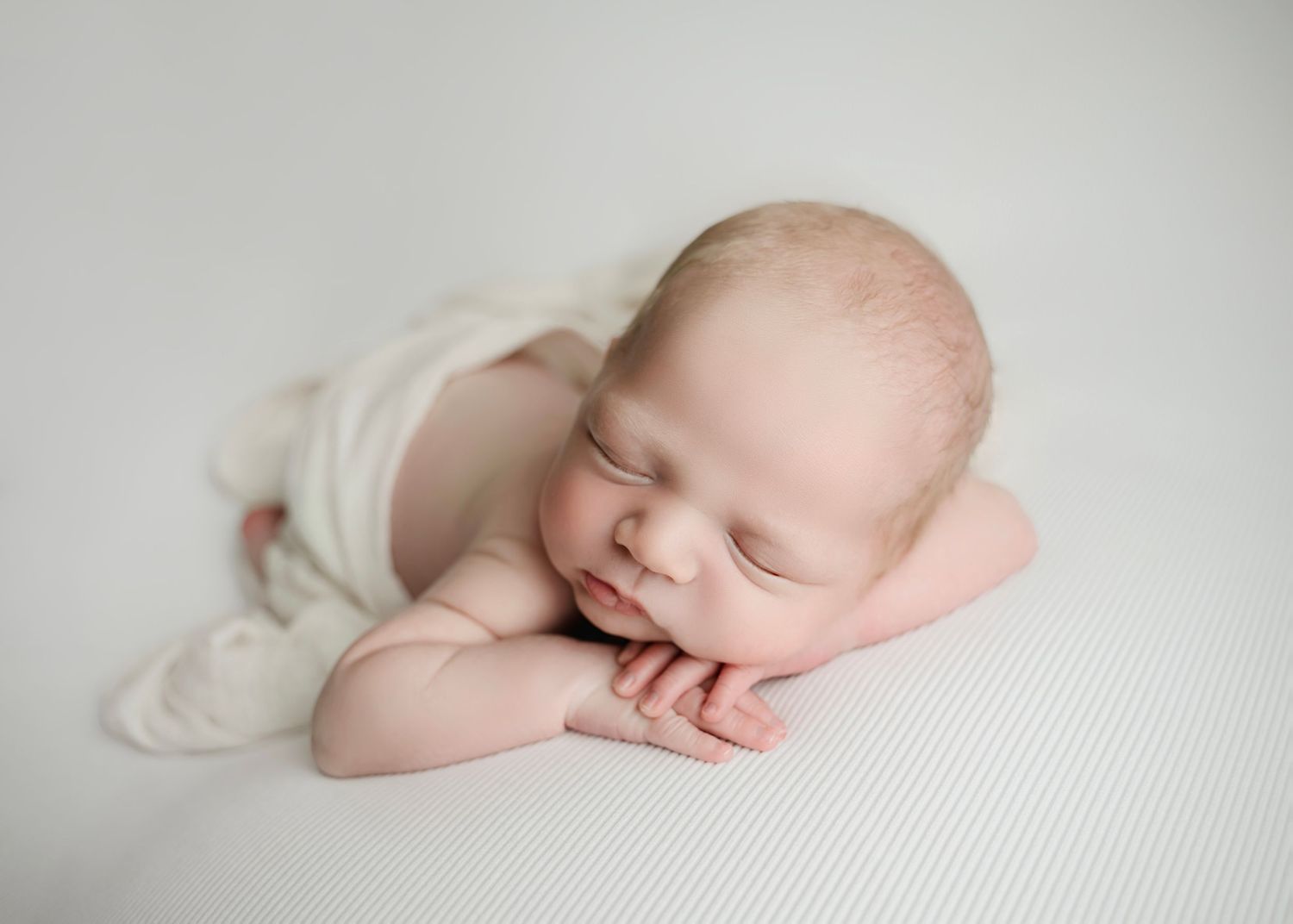 Peaceful newborn baby sleeping on white blanket, tiny hands tucked under chin in adorable posed newborn photography session.
