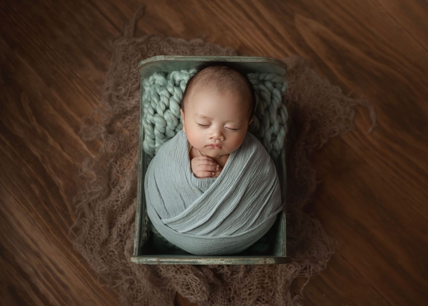 Sleeping newborn baby swaddled in sage green wrap, nestled in a rustic wooden box with chunky knit blanket.