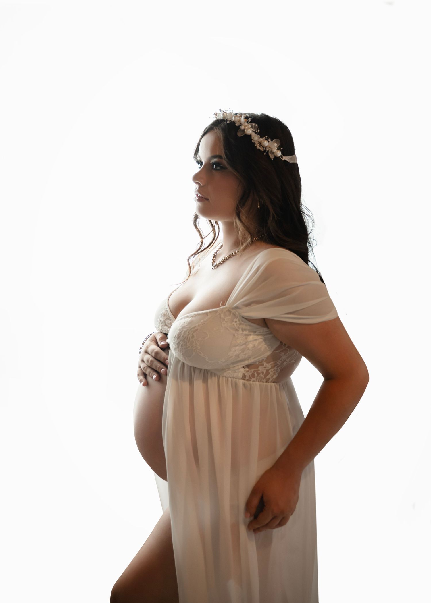 Pregnant woman in sheer ivory gown and floral crown poses gracefully against white background in elegant maternity portrait.