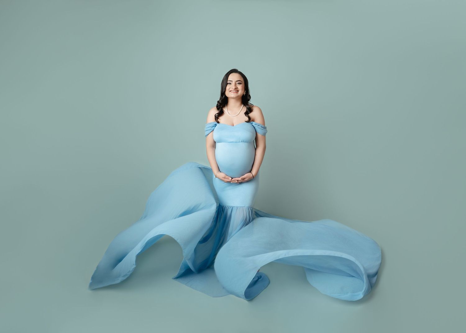 Pregnant woman in flowing light blue off-shoulder gown poses gracefully against a soft teal studio backdrop.