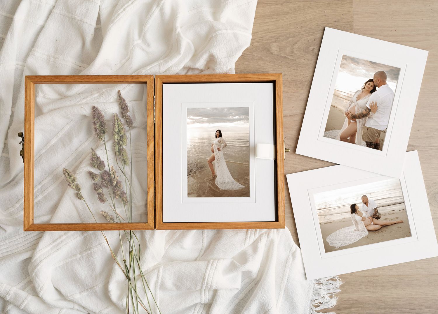 Wooden photo box with dried flowers beside framed beach wedding portraits on white linen background.