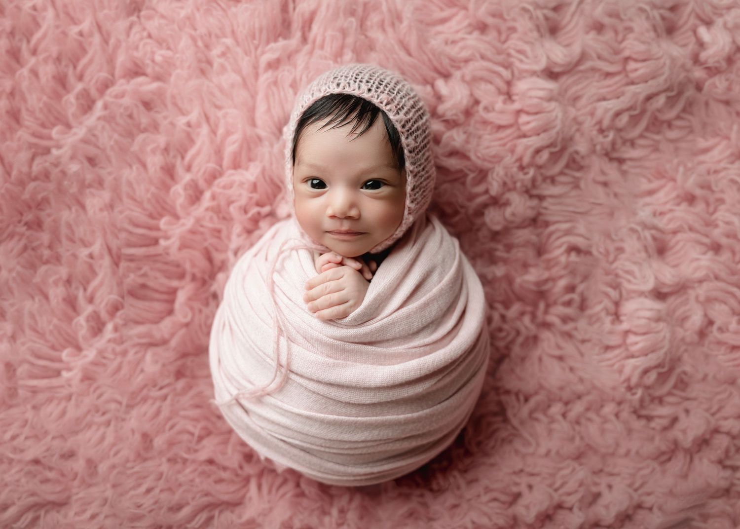Newborn baby swaddled in pink wrap wearing knit bonnet, posed on fluffy pink fur backdrop.