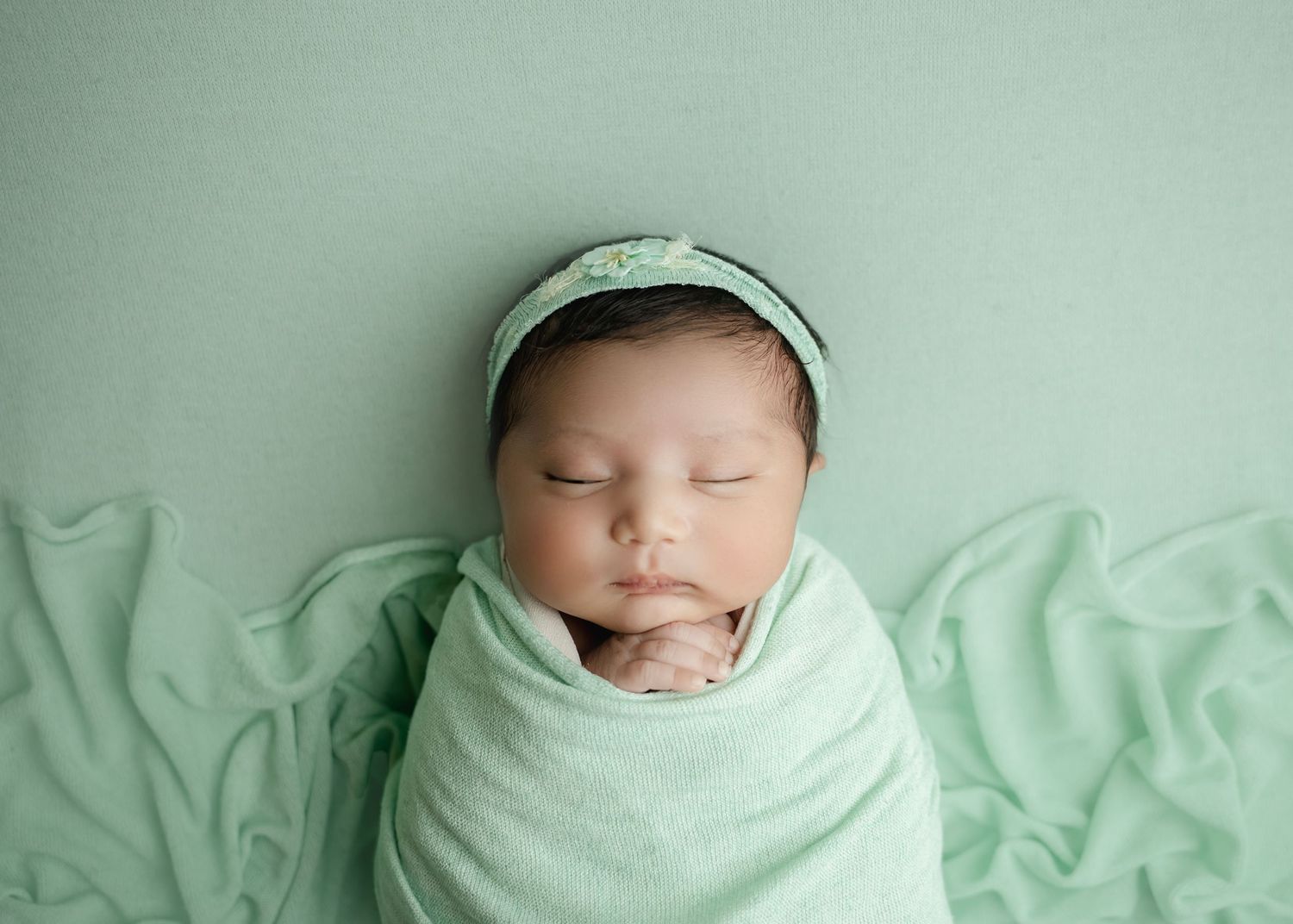 Newborn baby sleeping peacefully swaddled in a mint green wrap with matching headband on green background.