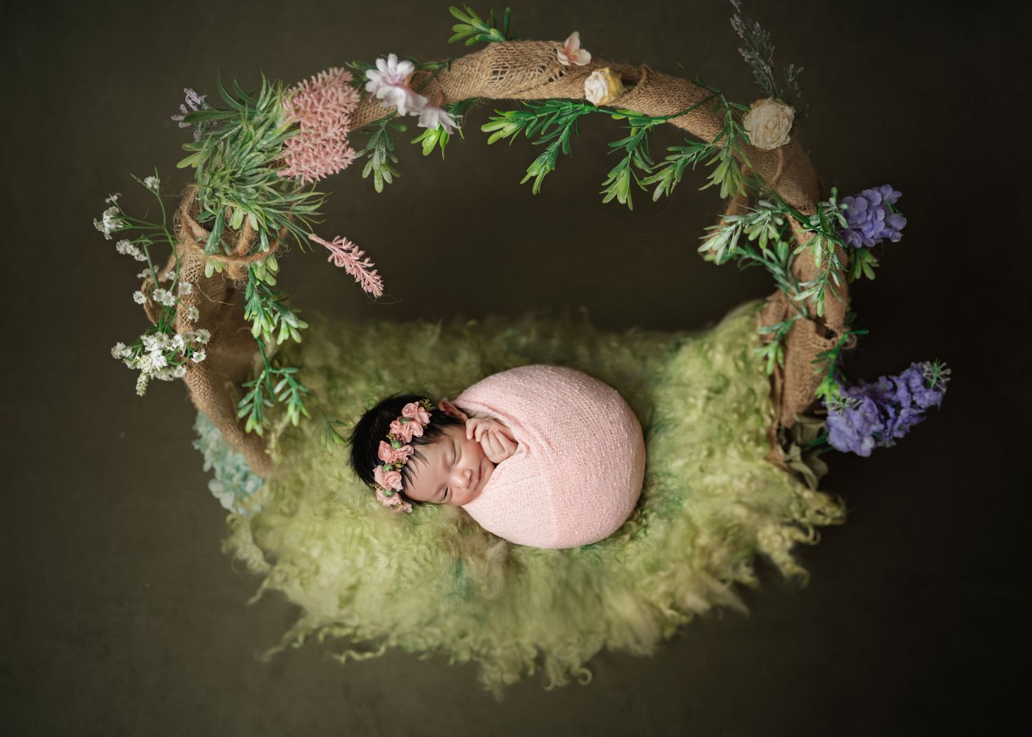 Newborn baby sleeping in a floral wreath nest with green moss and purple flowers in a professional photo.