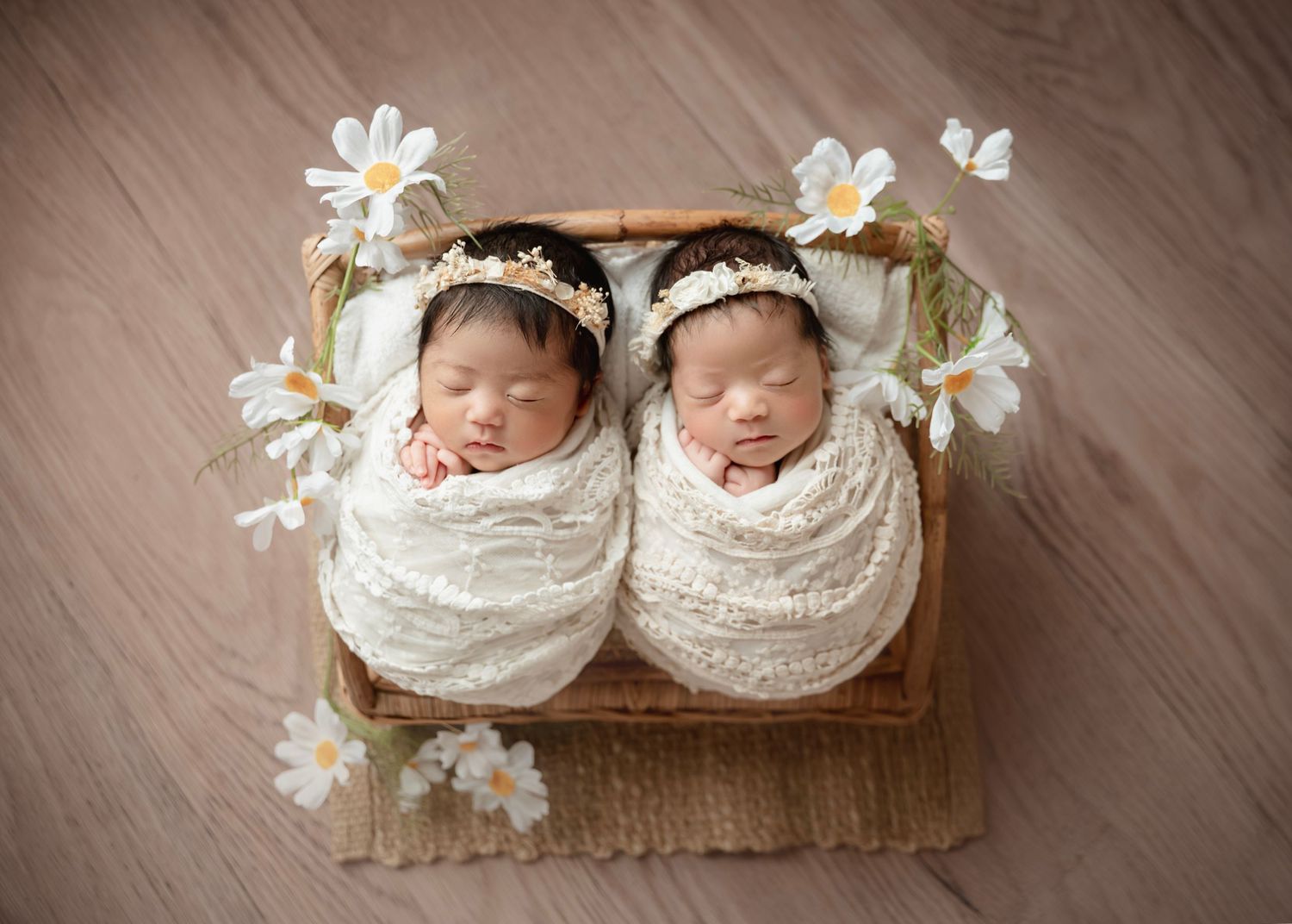 Two newborn twins swaddled in white lace wraps, wearing floral crowns, sleeping peacefully in a wooden basket with daisies.