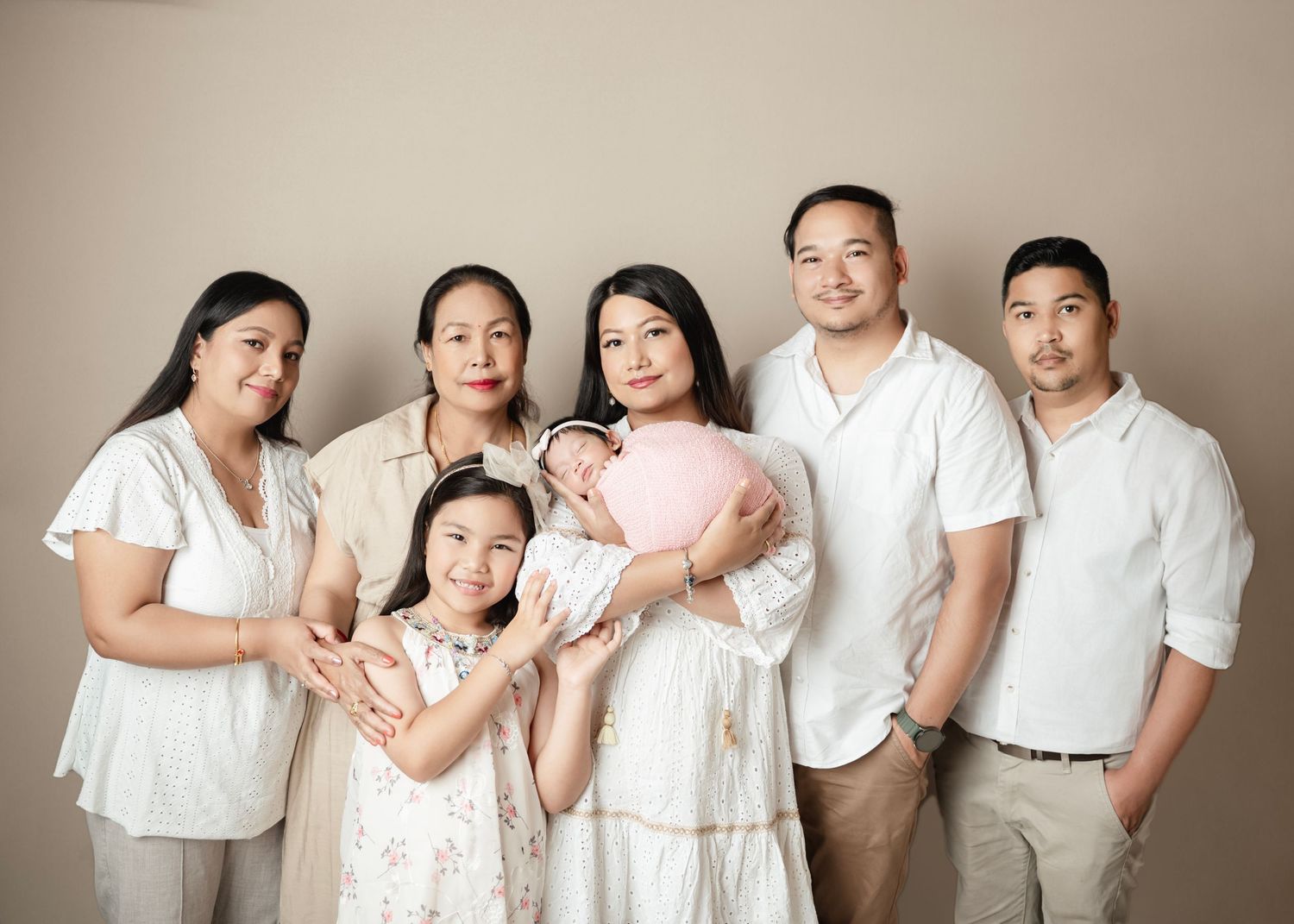 Multi-generational Asian family portrait in white outfits, featuring grandparents, parents, children, and a newborn baby in pink.