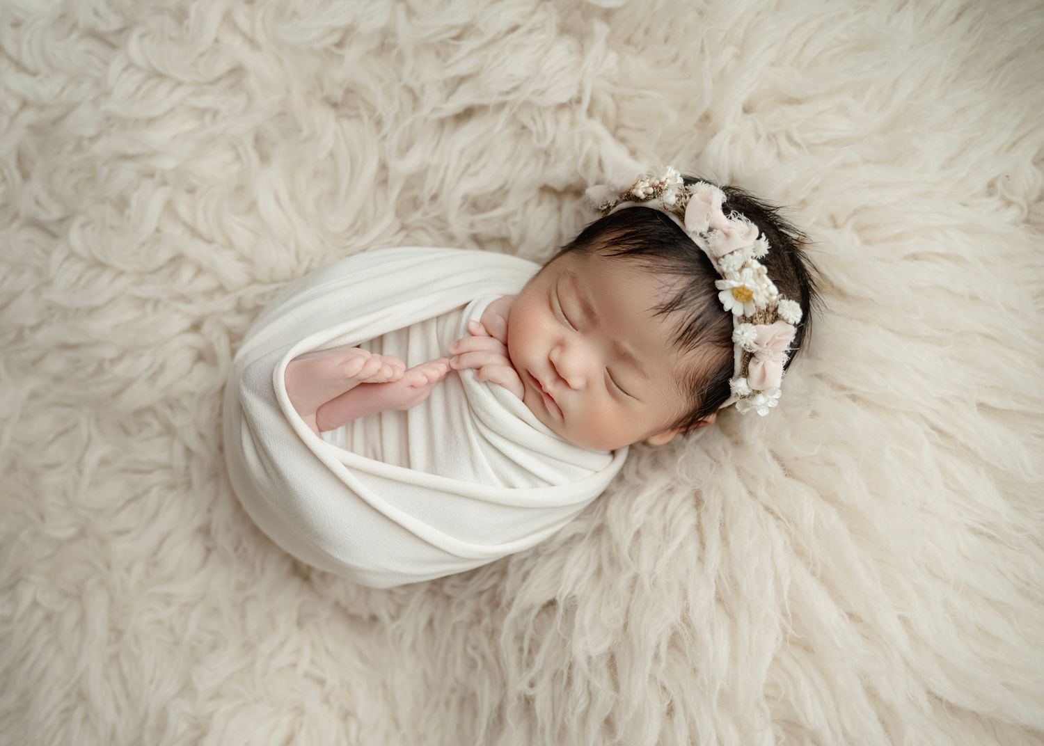 Sleeping newborn baby swaddled in white wrap with floral headband on cream fluffy rug.