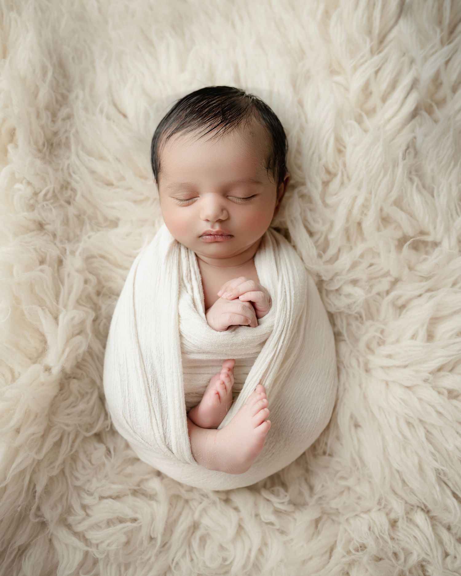 Peaceful newborn baby swaddled in cream wrap sleeping on fluffy white flokati rug in professional studio photo.