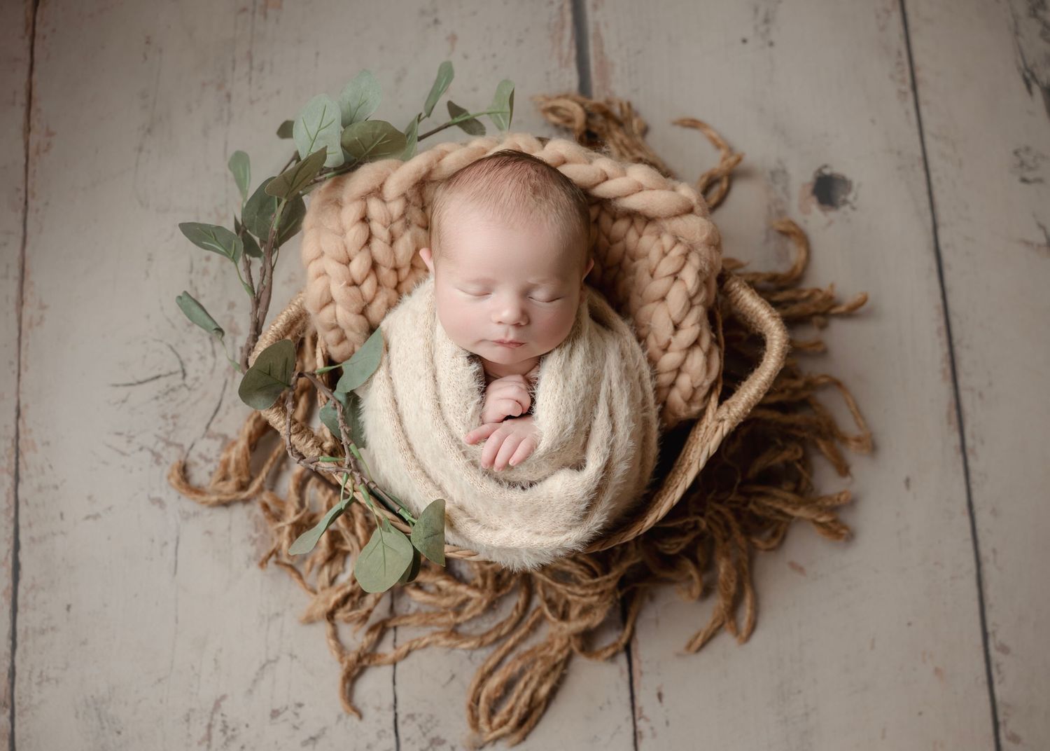 Newborn baby swaddled in cream wrap sleeping peacefully in a rustic basket with chunky knit and eucalyptus.