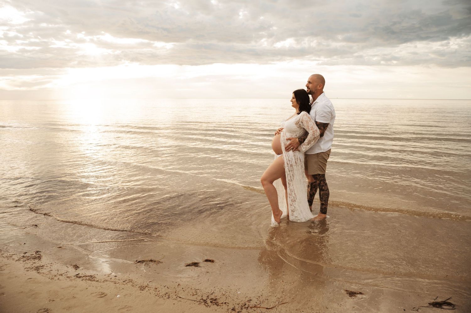 Pregnant couple embracing on a serene beach at sunset, waves reflecting golden light around their feet.