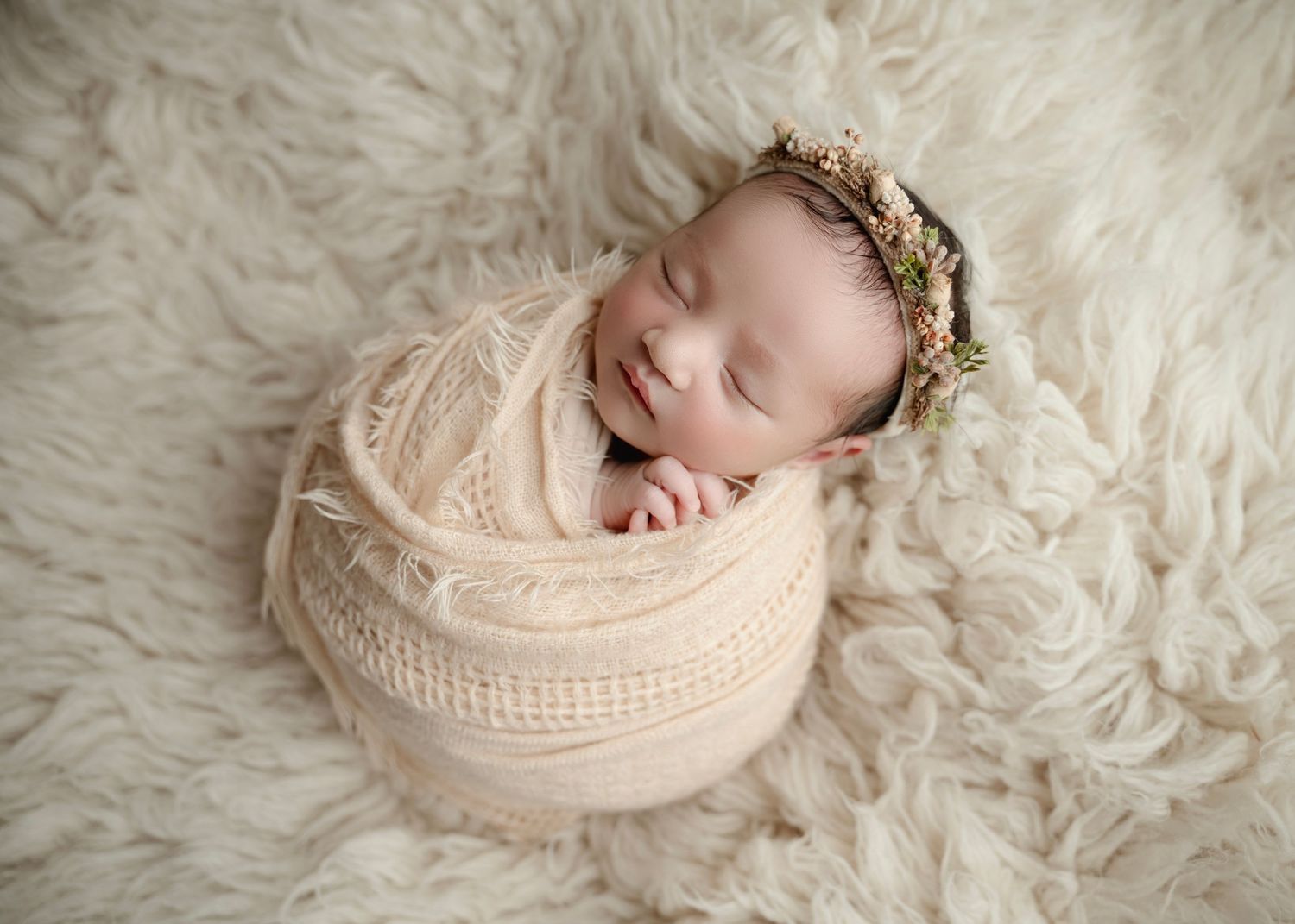Sleeping newborn baby wrapped in cream knit swaddle with floral crown on fluffy white fur rug.