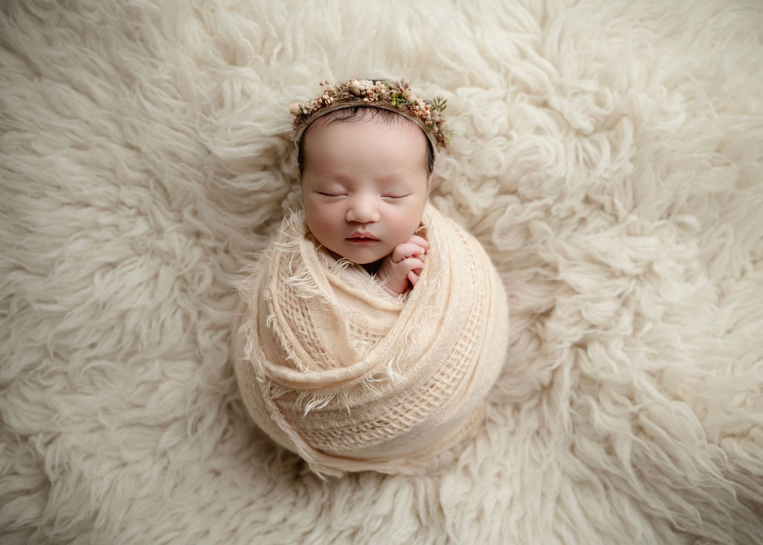 Sleeping newborn baby wrapped in cream knit swaddle with floral crown on fluffy white fur backdrop.