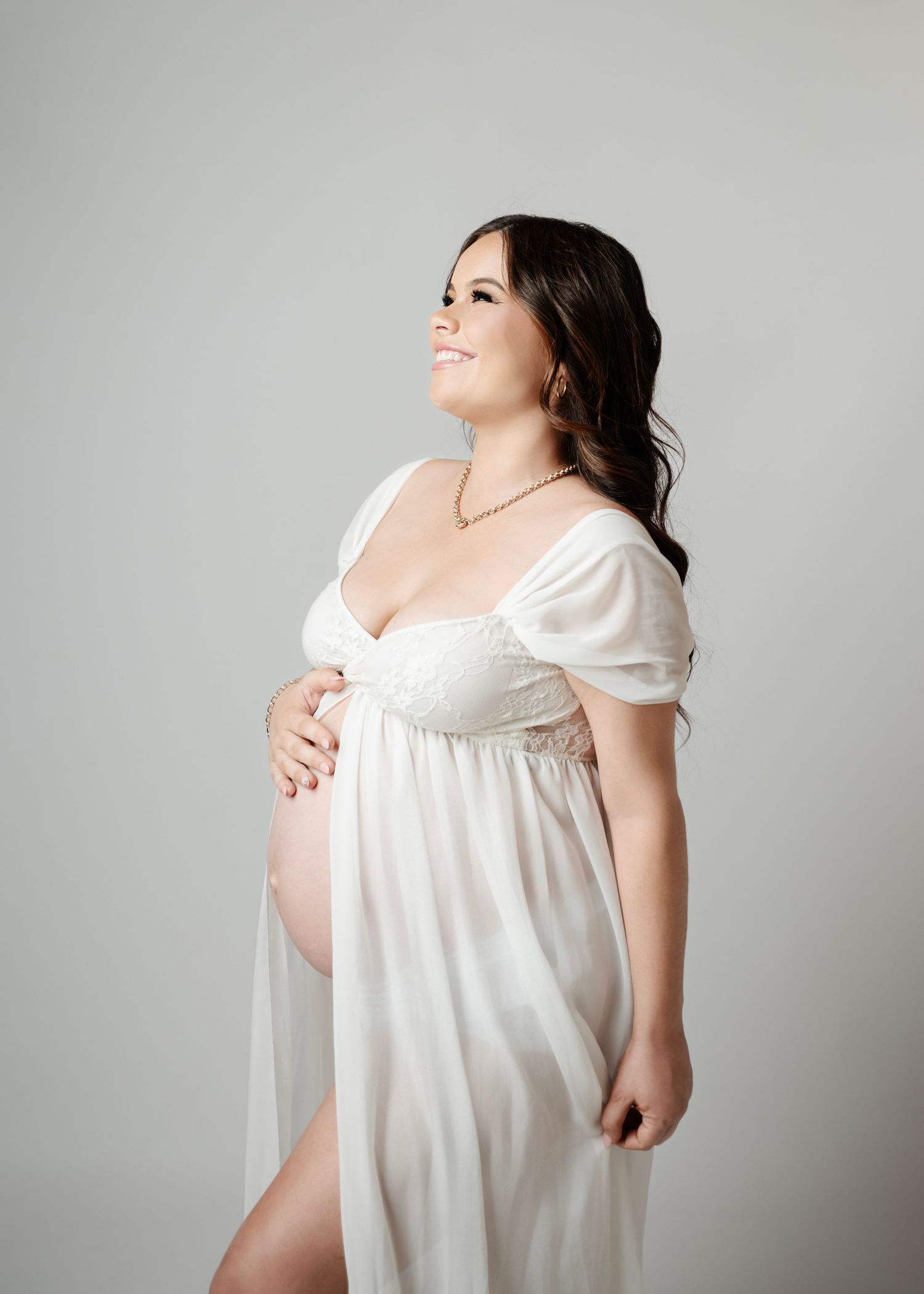 Smiling pregnant woman in white maternity gown cradling baby bump in a bright studio setting.