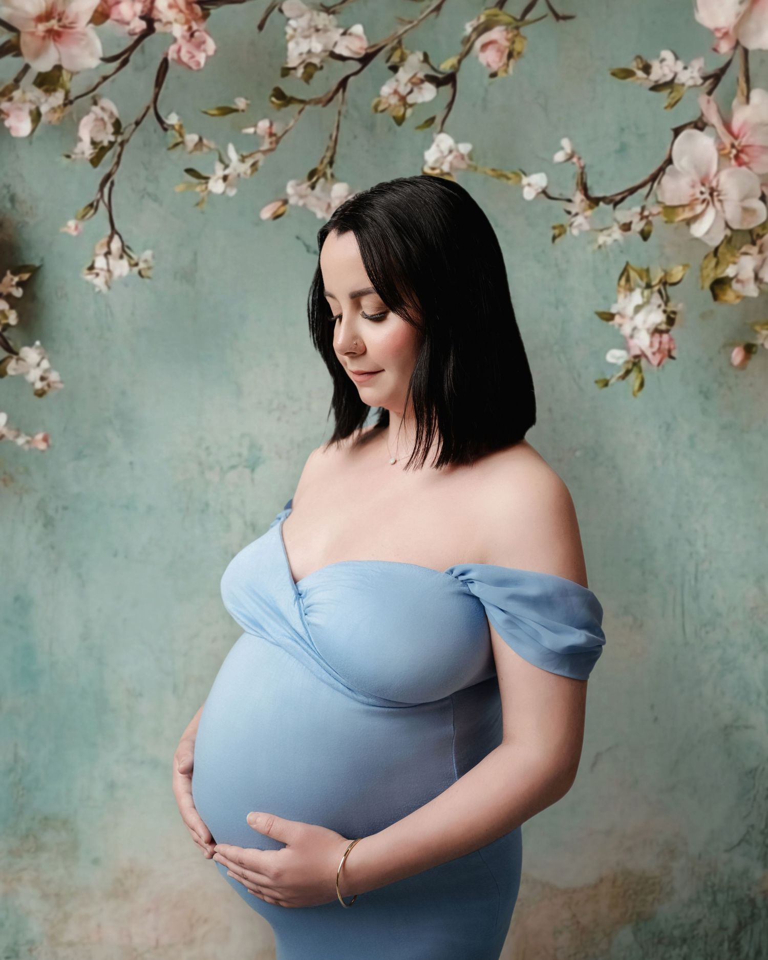Pregnant woman in blue off-shoulder gown holds baby bump, posing before a floral backdrop with cherry blossoms.