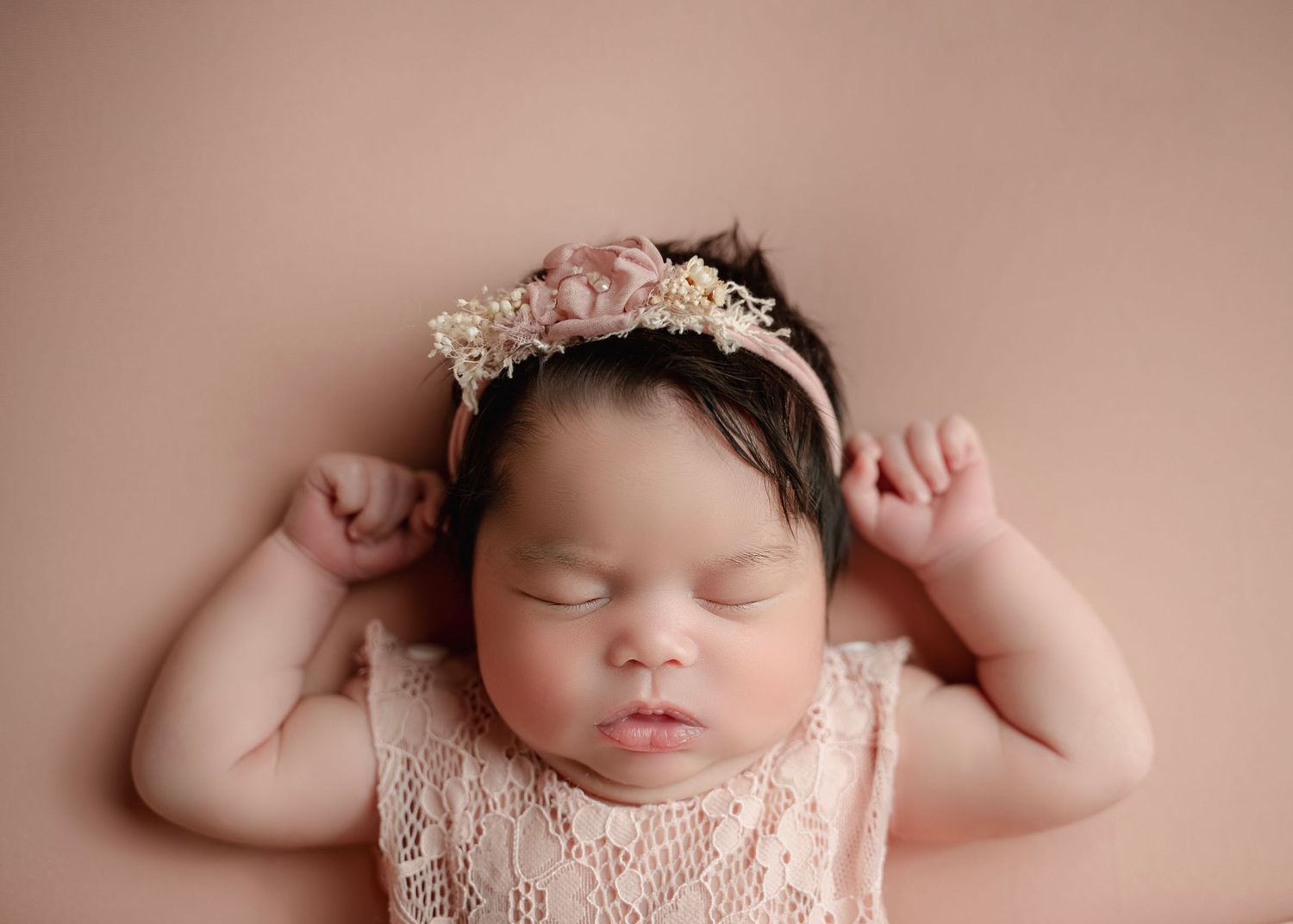 Sleeping newborn baby girl in pink lace outfit and floral headband on a soft pink backdrop.
