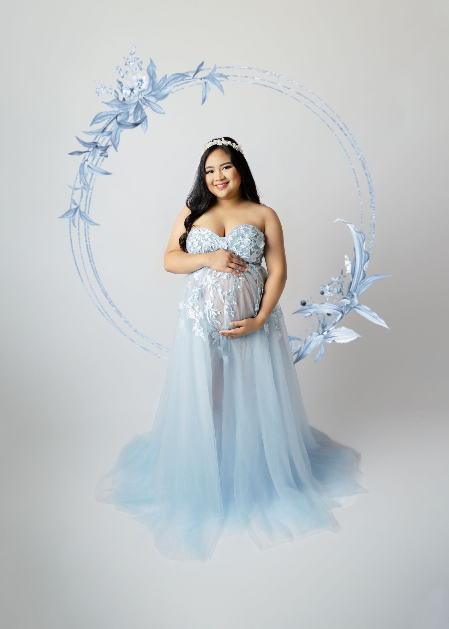Pregnant woman in light blue tulle maternity gown with floral crown poses gracefully in studio setting.
