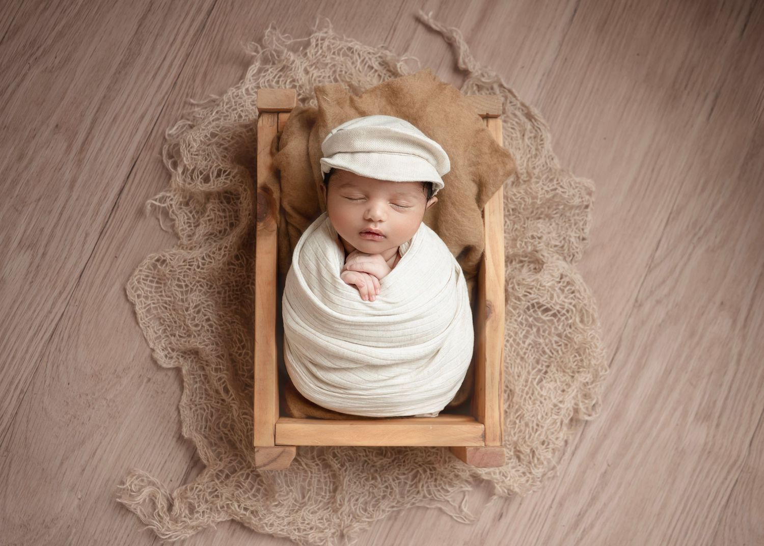 Newborn baby swaddled in white wrap wearing a newsboy cap, sleeping peacefully in a small wooden crate prop.
