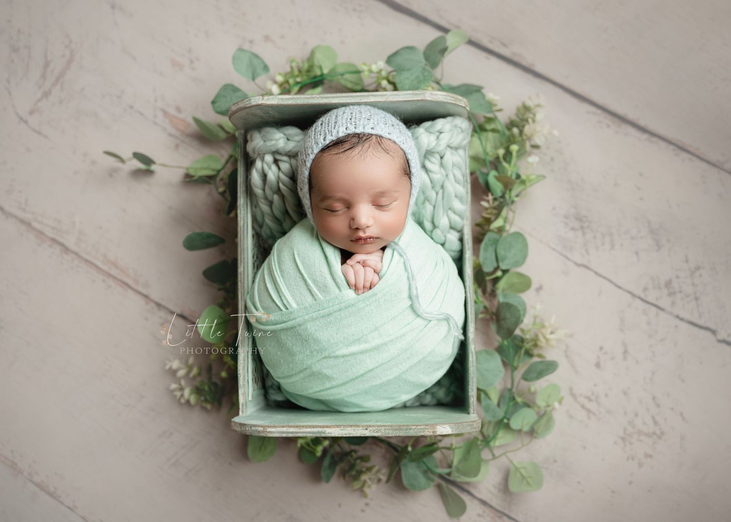 Newborn baby wrapped in mint green swaddle sleeping in a rustic wooden box surrounded by eucalyptus leaves.