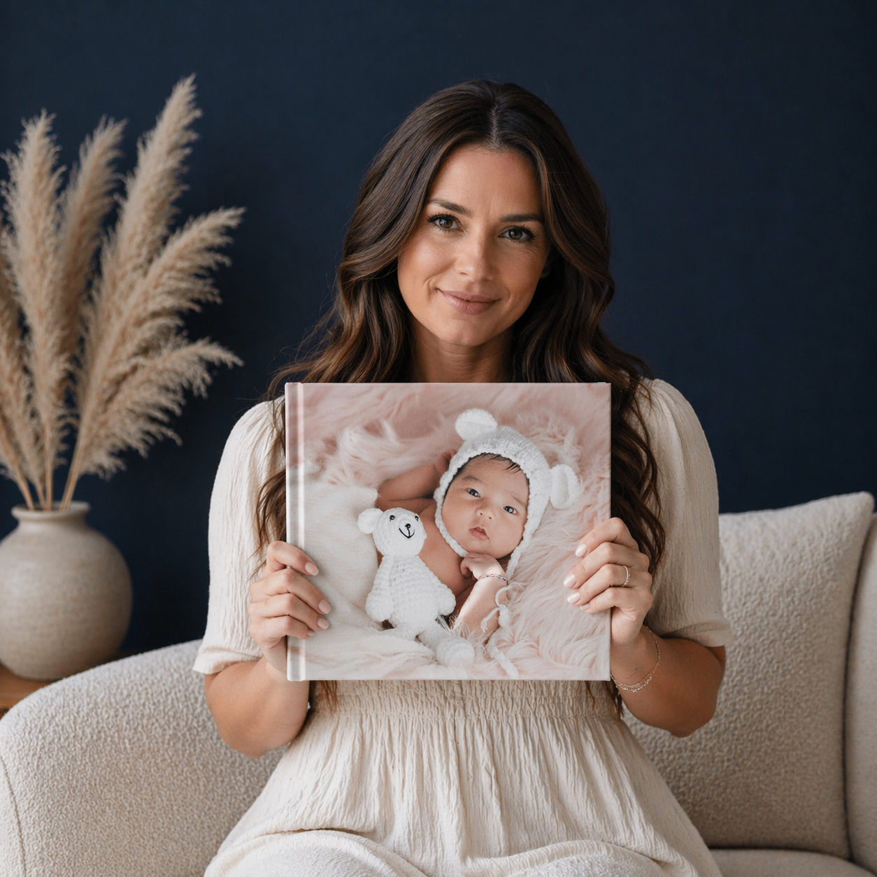 A woman in a white dress holds a photo book featuring a newborn baby wearing a bear hat, sitting in a cream chair.