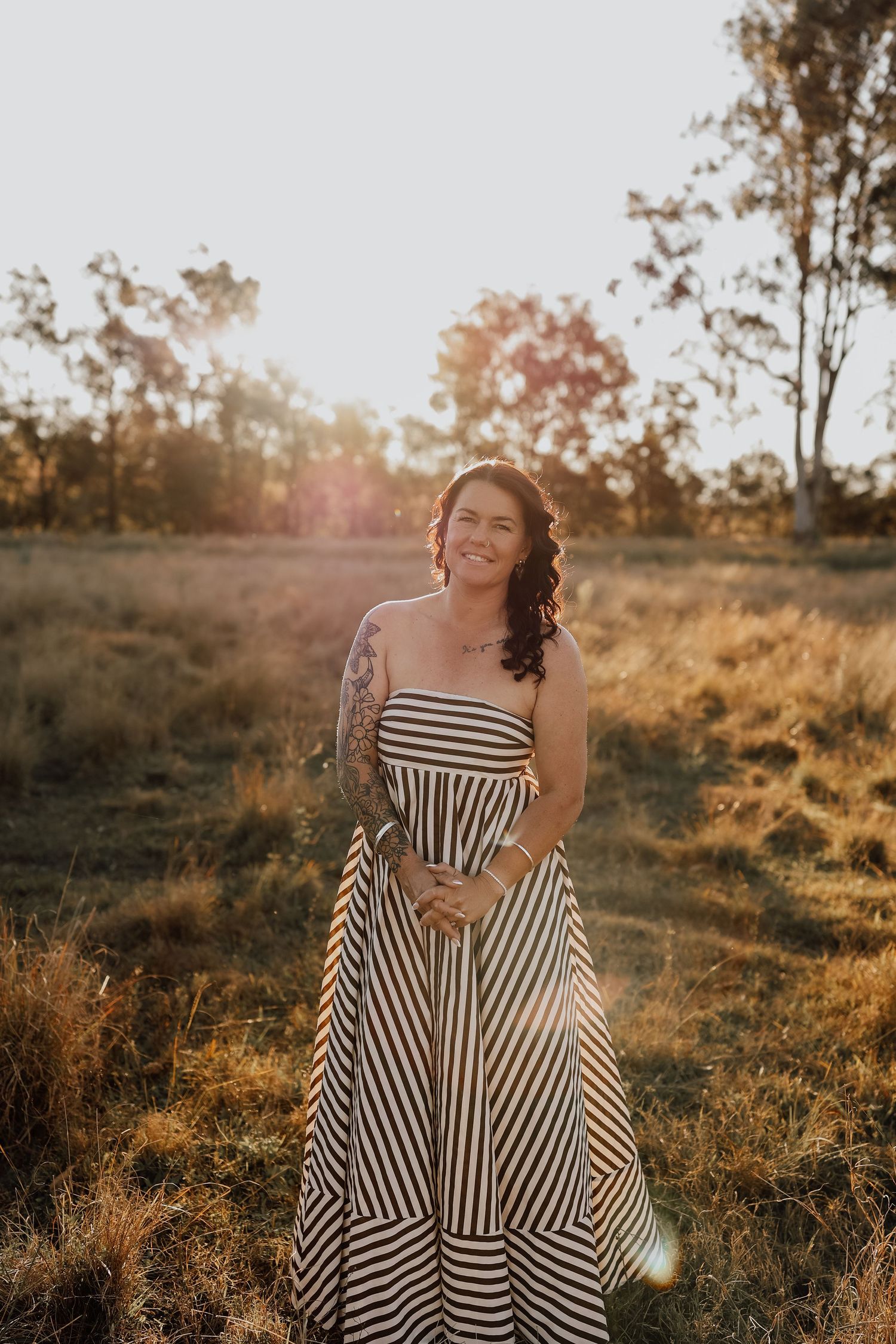 A striped maxi dress glows in golden sunlight against a backdrop of autumn trees and tall grass at sunset.