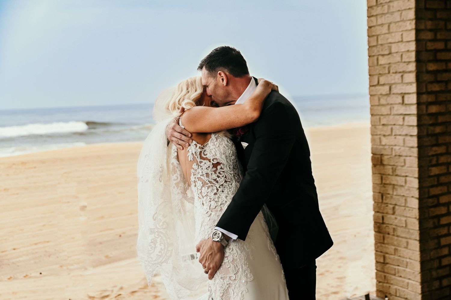 A romantic moment captured against a beige brick wall and beach backdrop.
