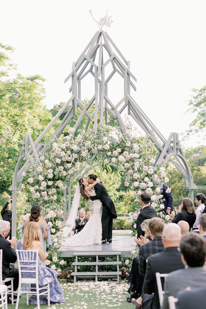 A romantic outdoor wedding ceremony takes place under a white gazebo adorned with pink roses and greenery.