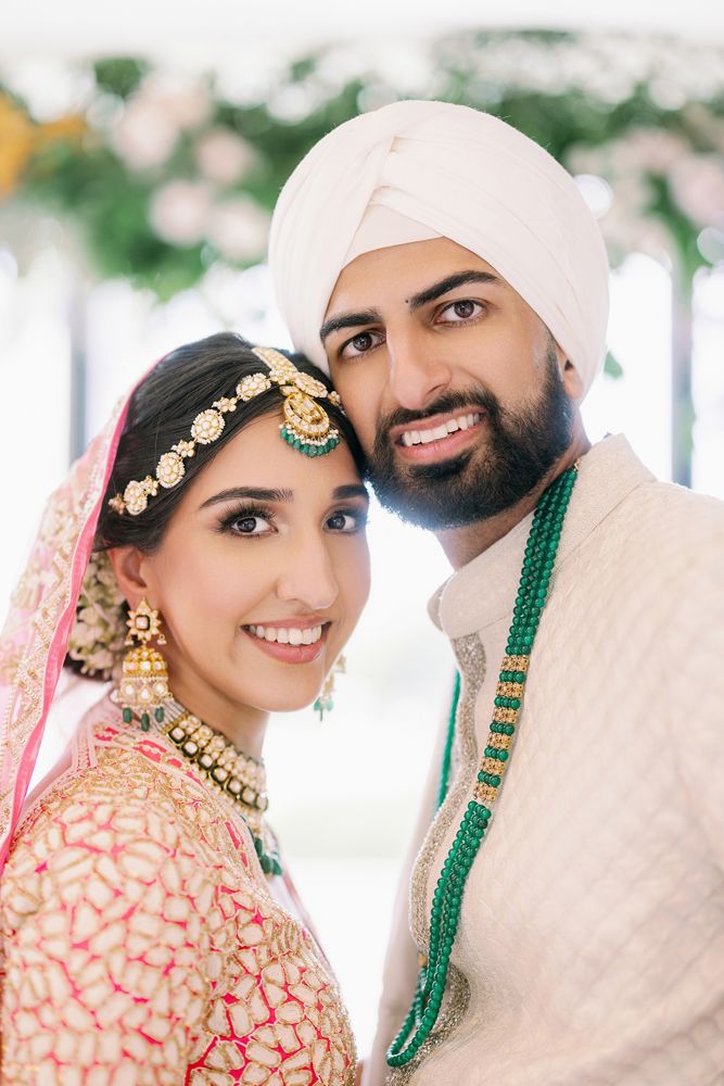 A couple in traditional South Asian wedding attire smiles together in a bright outdoor setting with blurred greenery.