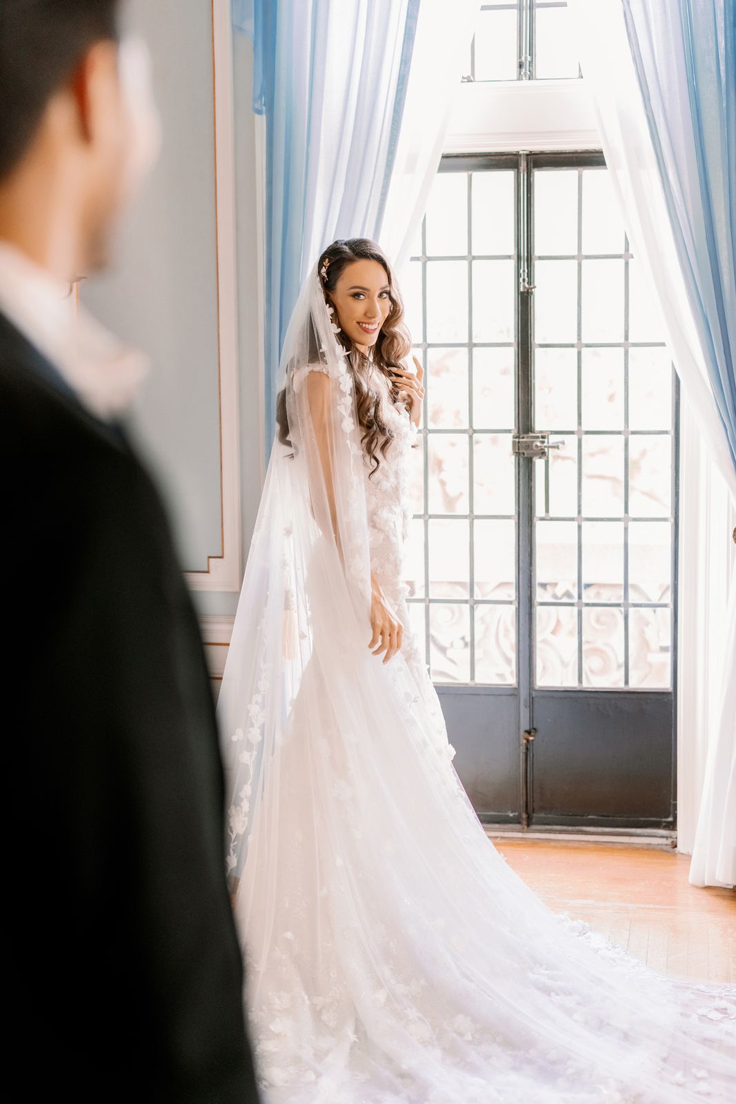 Bride in flowing white wedding gown stands gracefully by window in natural light.