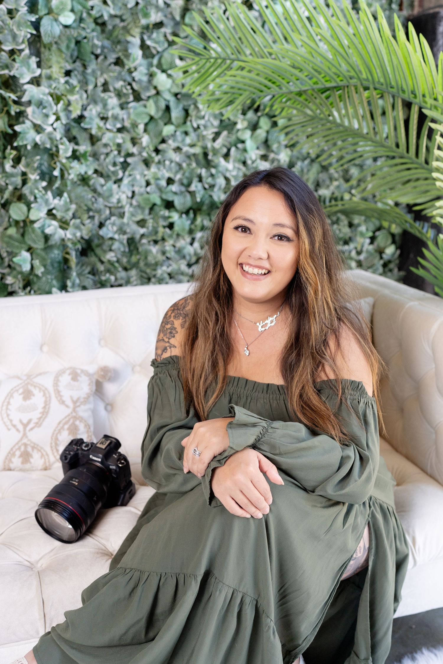 A photographer in an olive green off-shoulder dress sits on a cream couch with a camera nearby and tropical foliage behind.