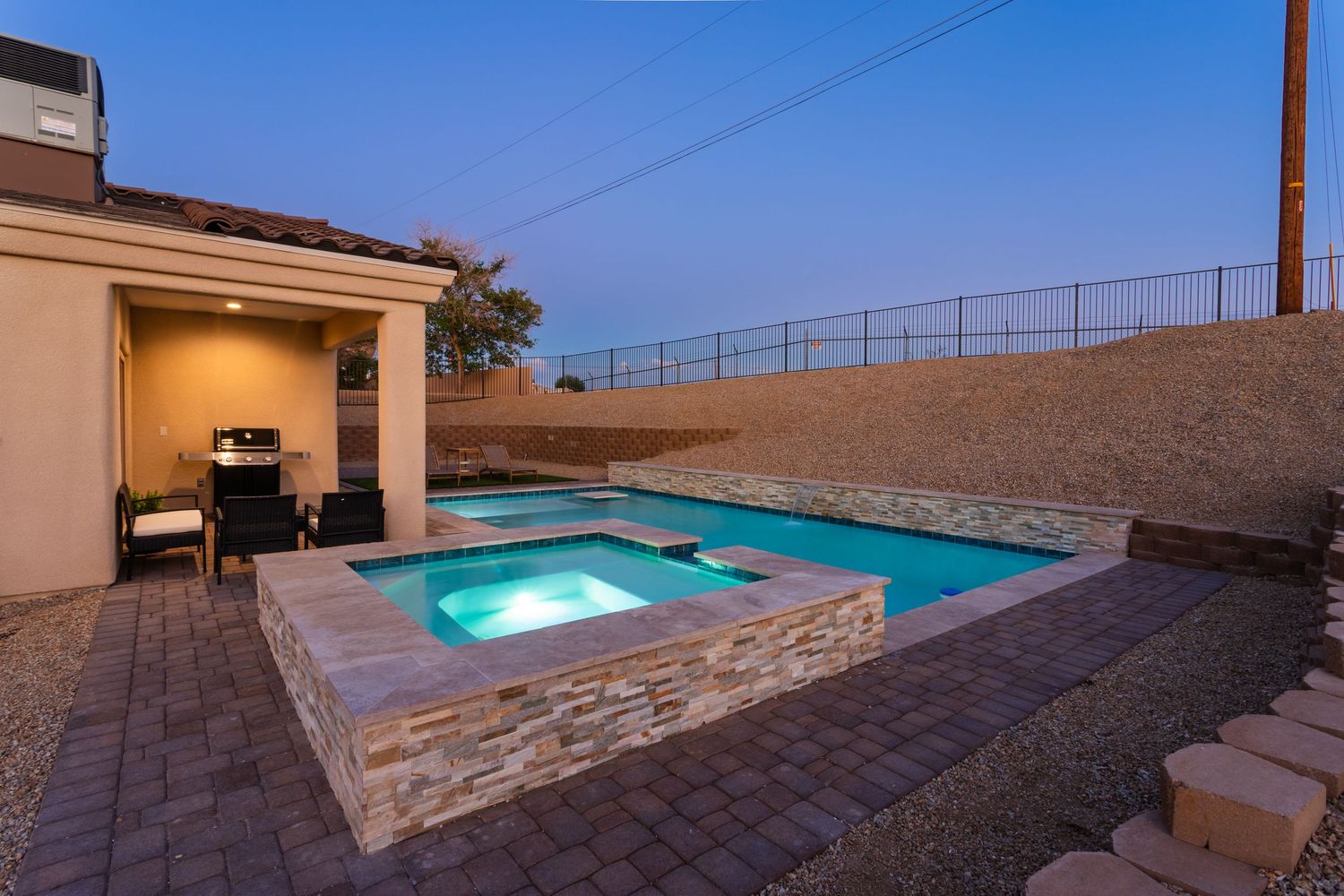 Illuminated stone hot tub with blue water glows at twilight against beige stucco house and desert landscape backdrop.