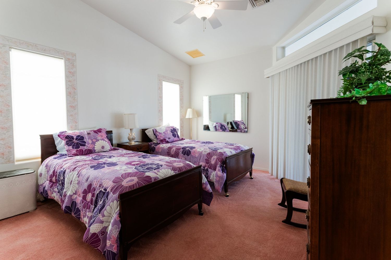 A spacious master bedroom with white walls and purple bedding features large windows and a ceiling fan for ventilation.