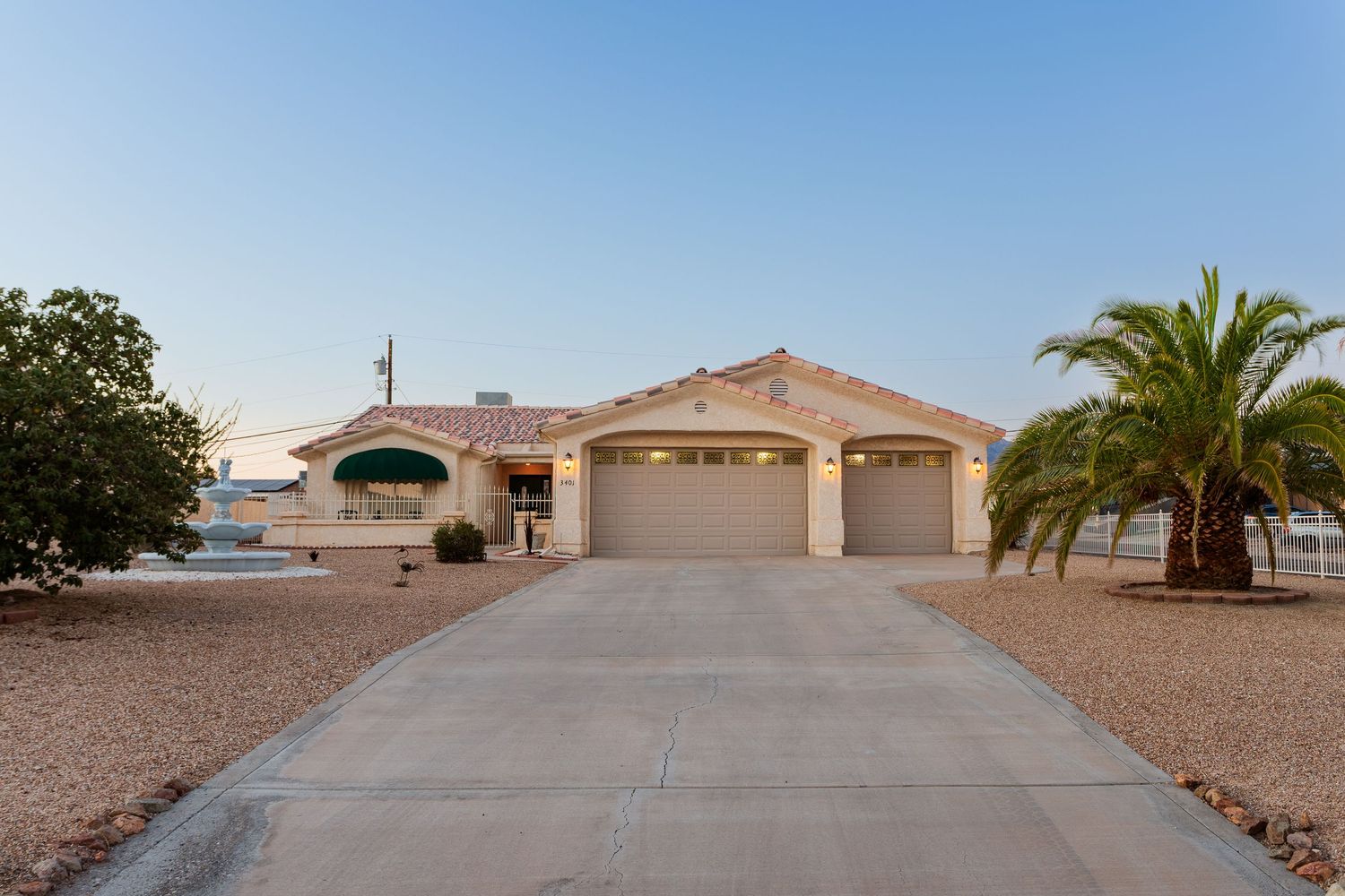 Tan desert home with three-car garage and palm tree landscaping in front of wide concrete driveway at dusk.