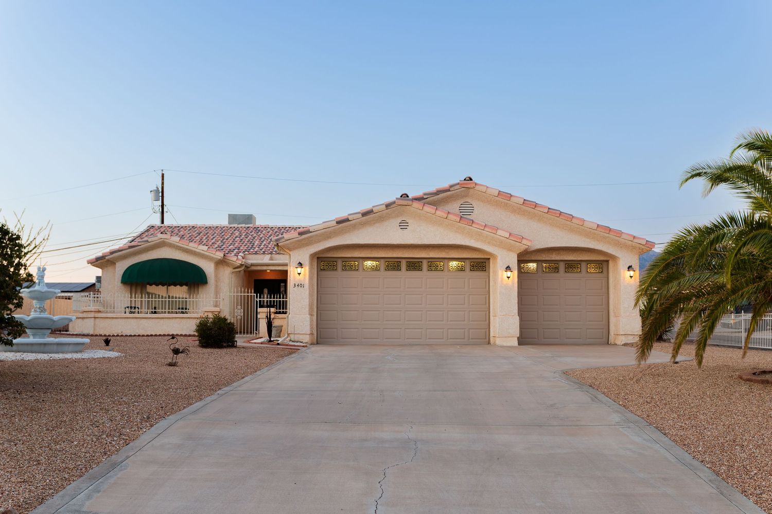 Desert home with a three-car garage and palm trees featuring beige stucco exterior and tile roofing at dusk.