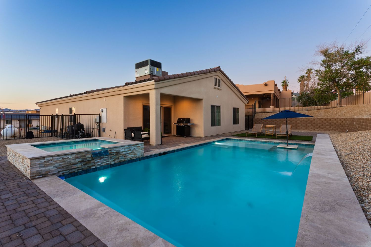 Large rectangular swimming pool with crystal blue water surrounded by desert landscape and residential buildings at sunset.