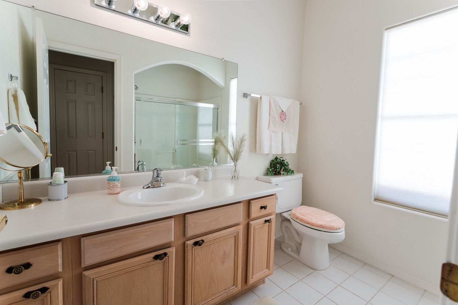 A modern bathroom interior with light wood cabinets, white countertops, and a large framed mirror above dual sinks.