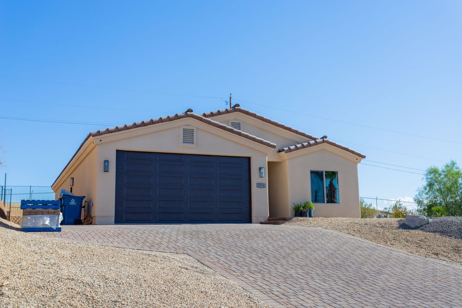 Modern single story stucco home with black garage door and paver driveway in desert landscaping setting.