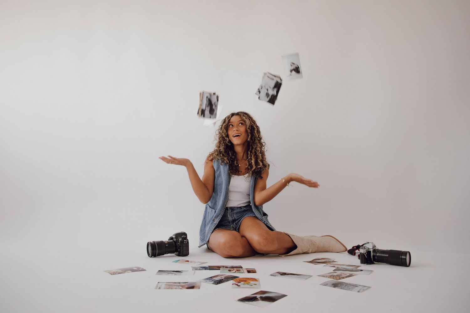 A photographer sits on the floor surrounded by scattered photographs and gestures with their hands.