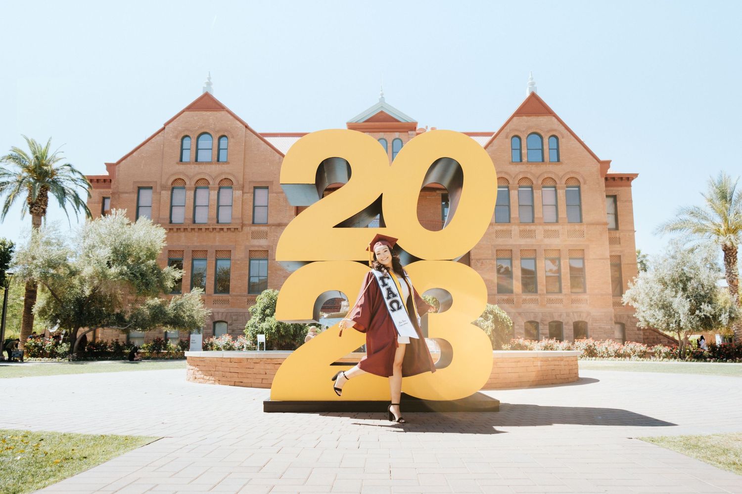 Graduate poses with large gold 2023 numbers in front of historic ASU building.