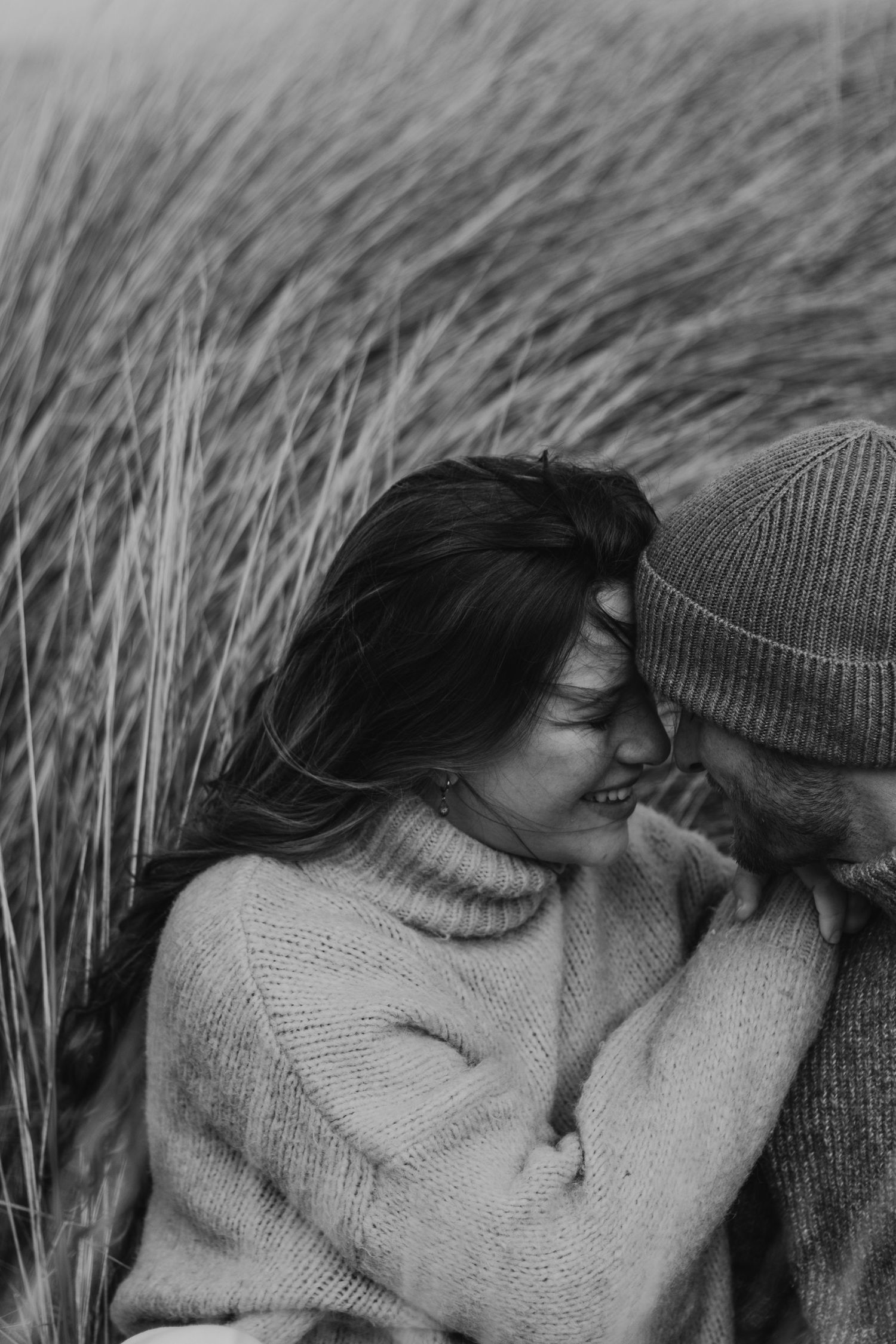 Black and white romantic photo of couple in sweaters embracing among tall grass in an intimate moment.