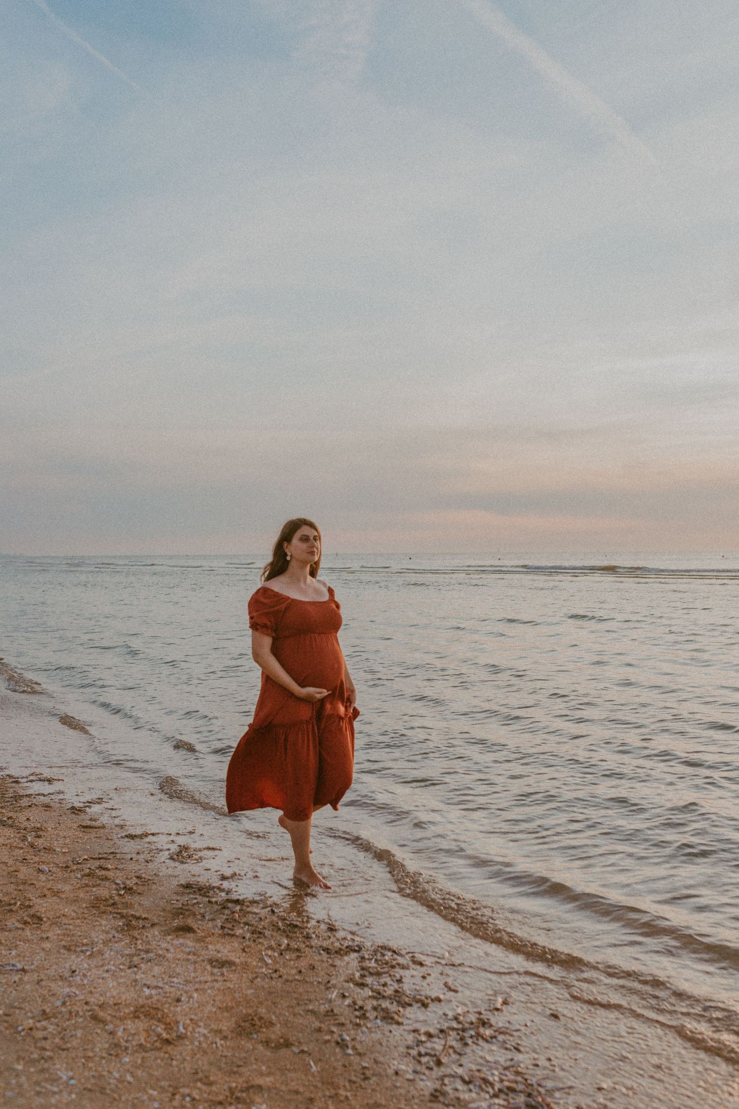 Person in orange dress walking along a serene beach shoreline at sunset with waves gently rolling in.