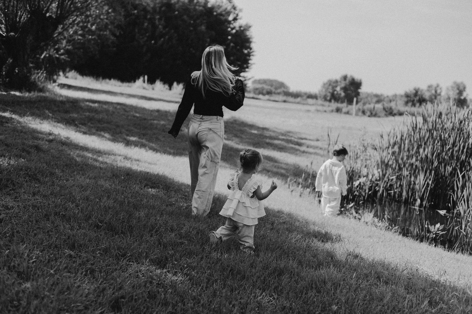 Black and white photo of a mother walking with two young children along a grassy path near a pond.