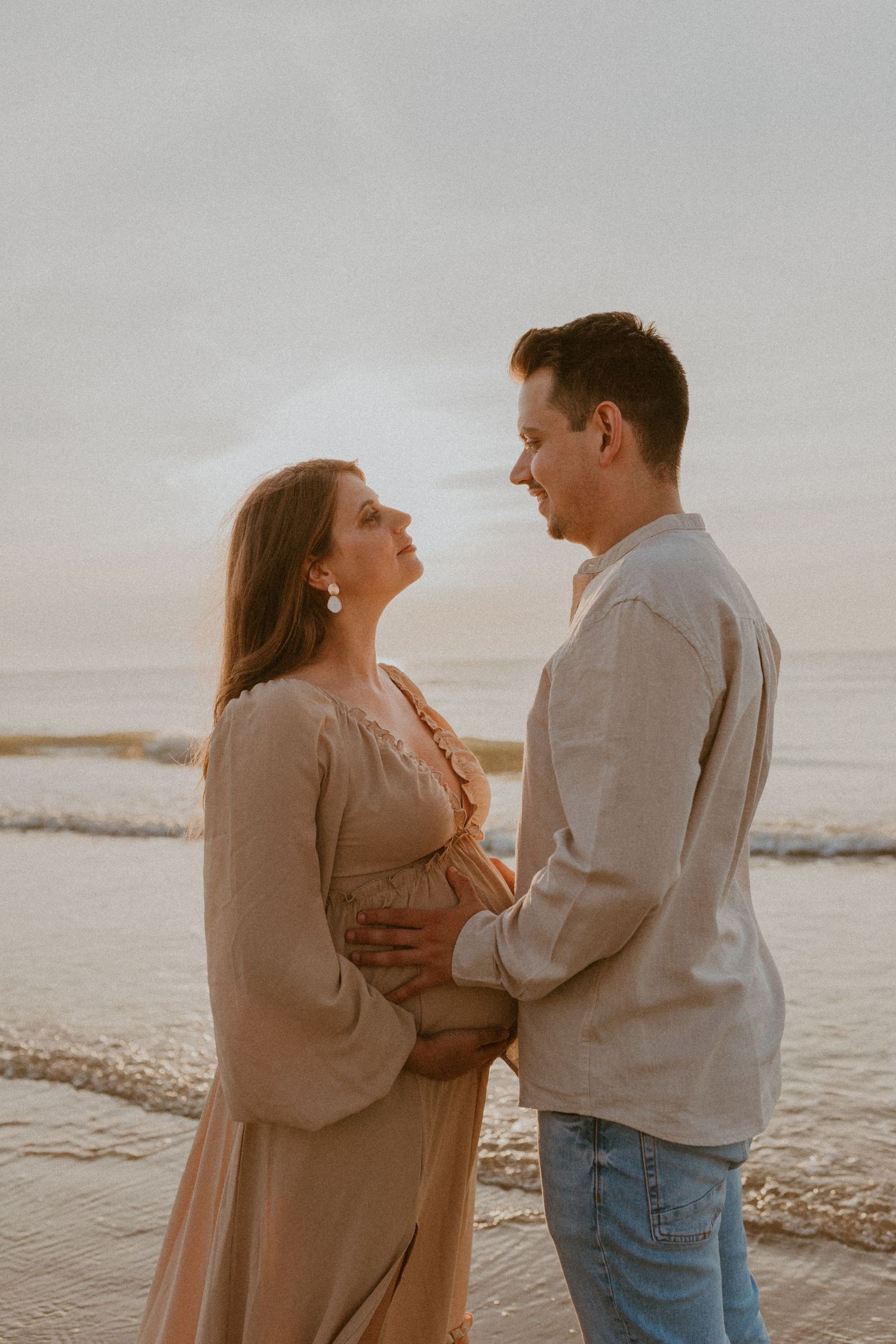 A couple shares an intimate moment on the beach at sunset wearing casual attire in warm muted tones.
