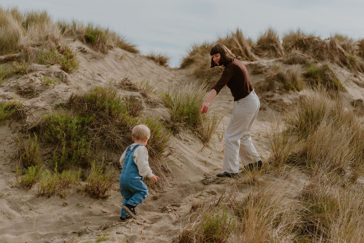 A mother and toddler climb sandy dunes with beach grass on a cloudy day.