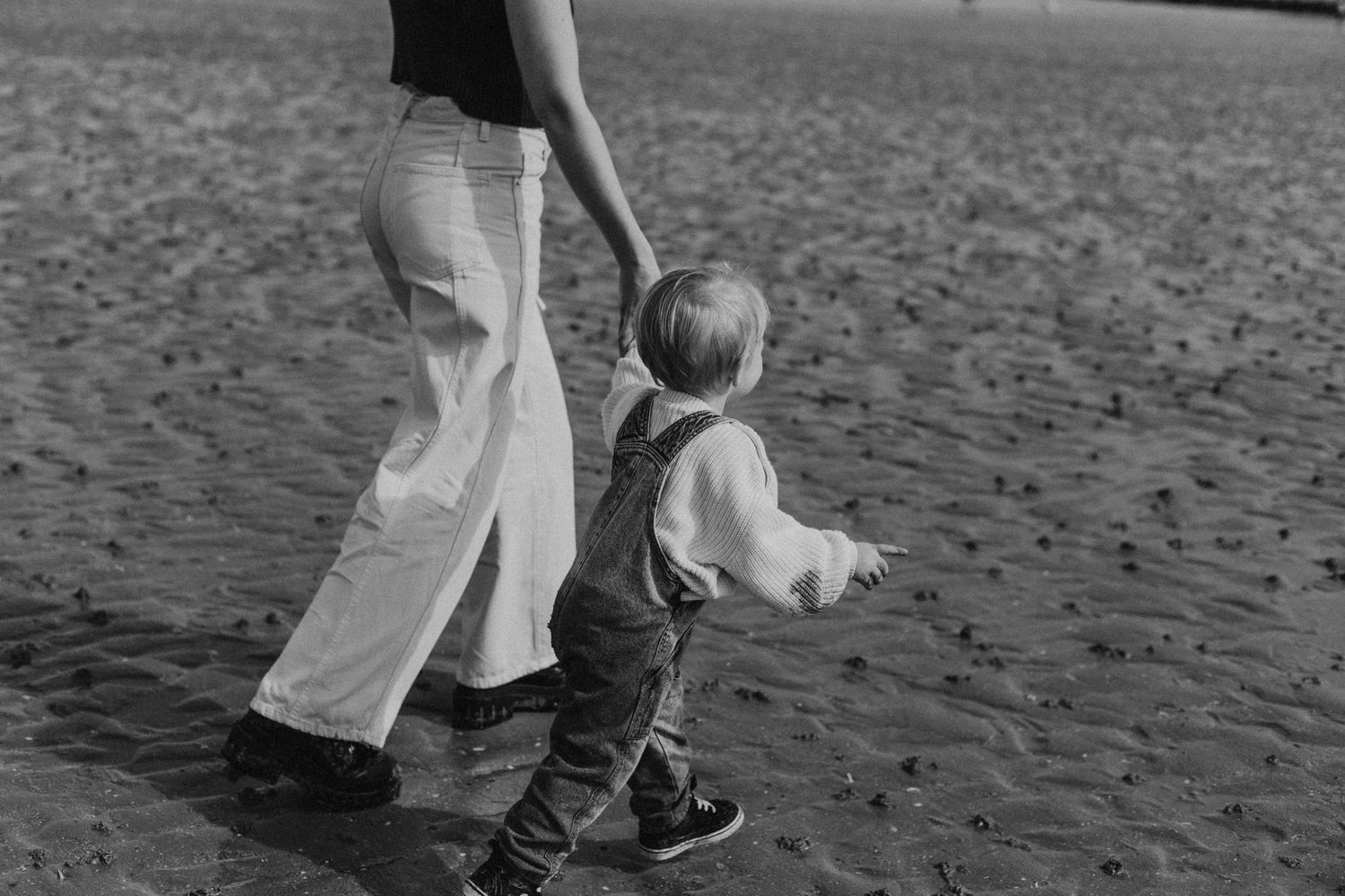 Black and white photo of a child holding an adult's hand while walking on a sandy beach.