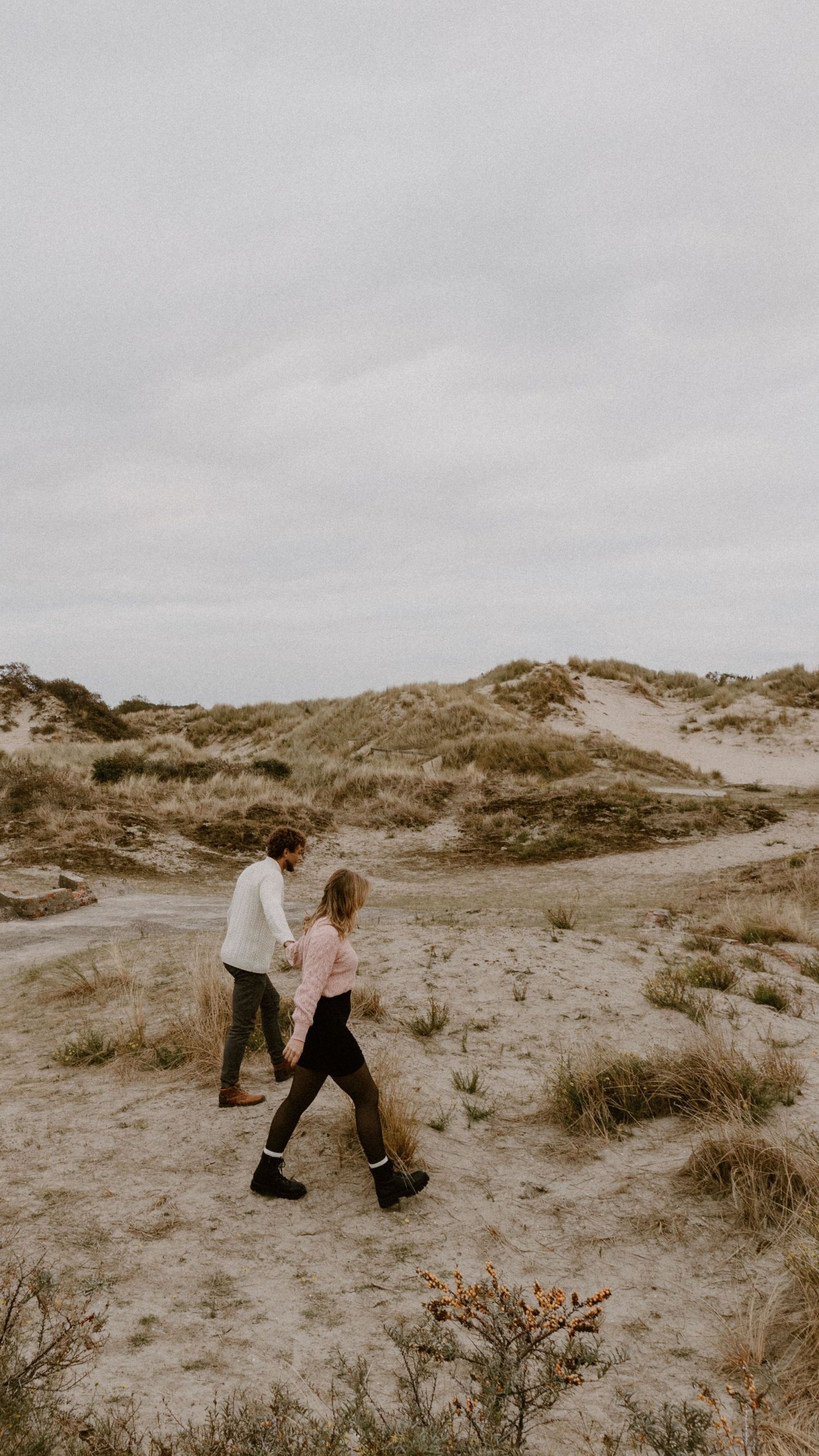 Twee mensen wandelen samen door zandduinen onder bewolkte luchten.