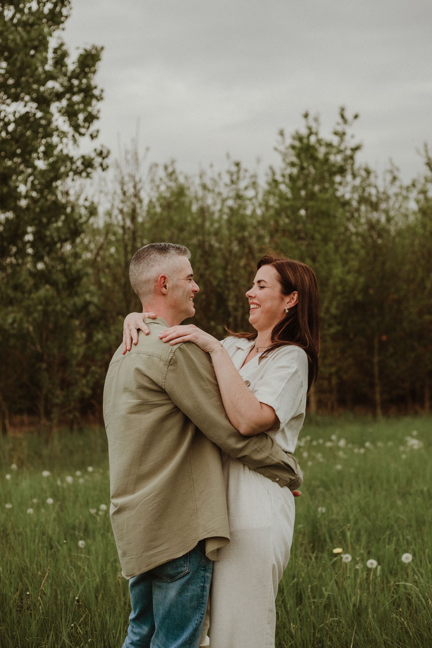 A couple embraces playfully in a meadow with wildflowers and trees in the background during a casual photoshoot.