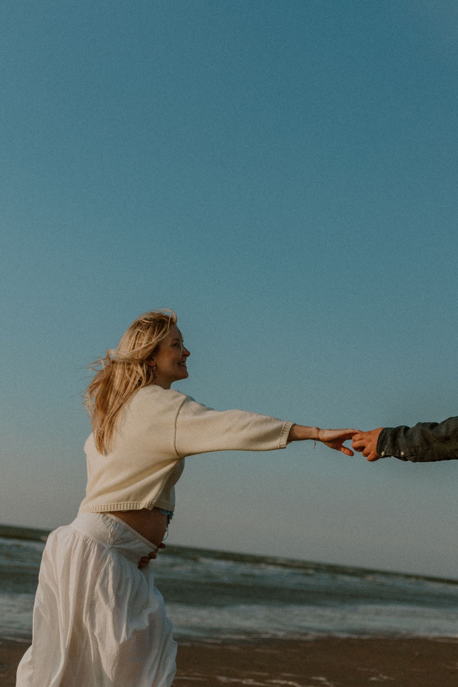 A blonde woman in a white sweater and skirt reaches out to hold hands on a breezy beach at sunset.